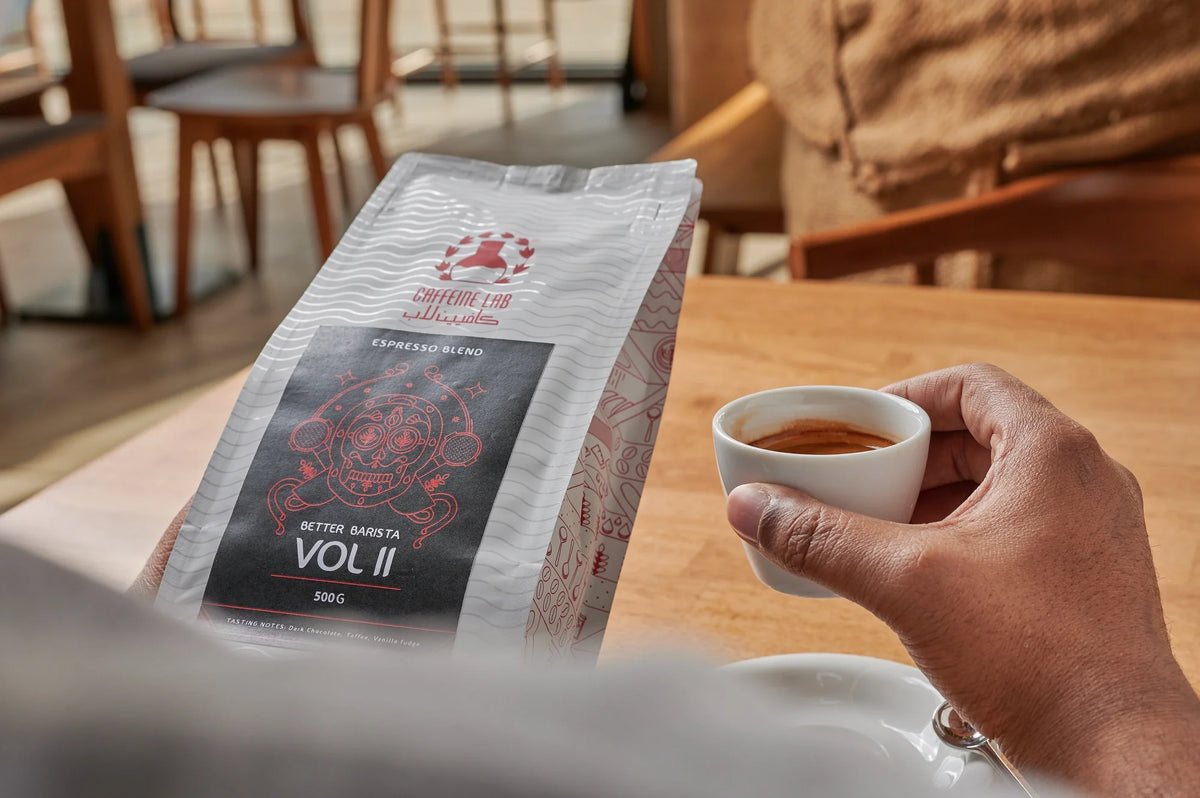 a coffee drinker holding a coffee bean bag of the better barista on the left hand and an espresso made by the same beans in the right hand and he is sitting on one of the tables of Caffeine Lab Café